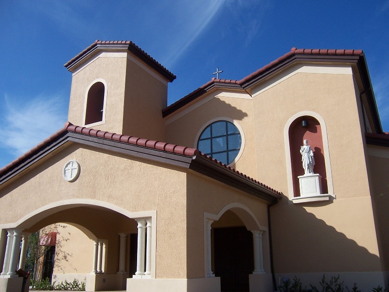 Front entrance of Our Lady Queen of Apostles church with its bell tower, statue niche and terracotta roof under a bright blue Florida sky